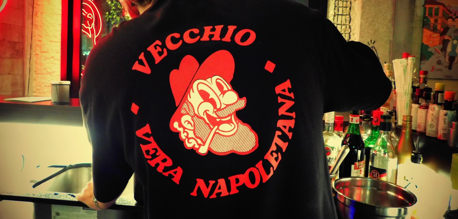 Emblema da casa, “Vecchio Vera Napoletana”, estampado em vermelho nas costas do uniforme, no salão em luz vermelha.